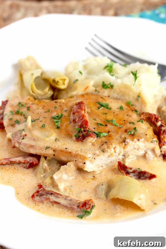 Creamy Sun-Dried Tomato Chicken Skillet Recipe 8 A piece of creamy chicken with artichoke hearts and sundried tomatoes on a white plate.