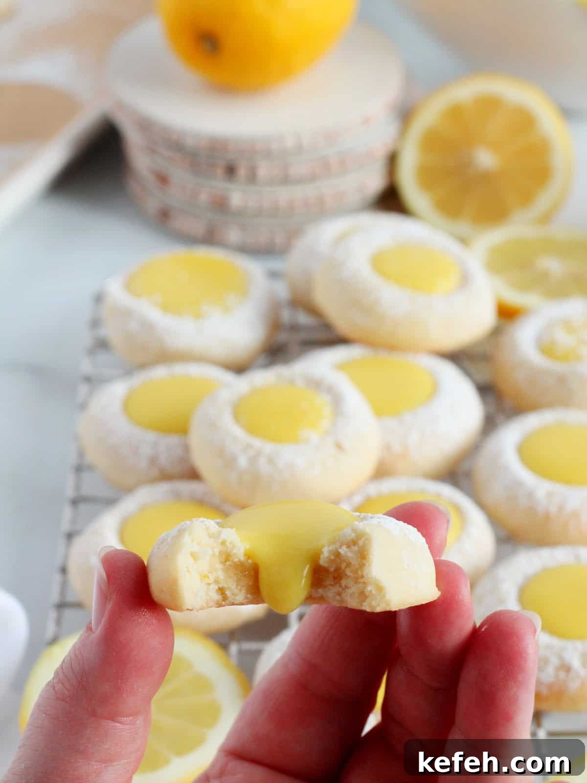 Iresistable lemon thumbprint cookie filled with homemamde lemon curd cut in half an placed in human hand. The lemon curd is slightly flowing down from the center. Plenty of lemon thumbpring cookies in the background with white decor and yellow lemons.