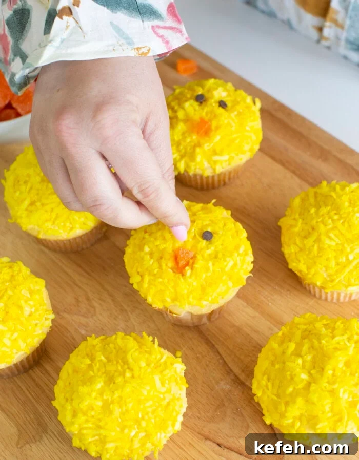 Process photo of making the Easter chick cupcakes face with mini chocolate chips for the 2 eyes and orange candy for the beak.
