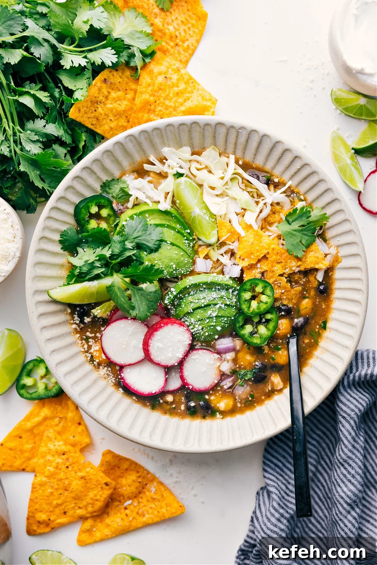 Quick 20-Minute Pozole Recipe Chicken Pozole served in a bowl topped with shredded cabbage, tortilla chips, avocado slices, radishes, cilantro, lime wedges, and jalapeño.