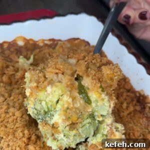 A close-up of a hand scooping a serving of Broccoli Cheese Casserole 🥦, topped with a golden brown crumb crust, from a white baking dish.