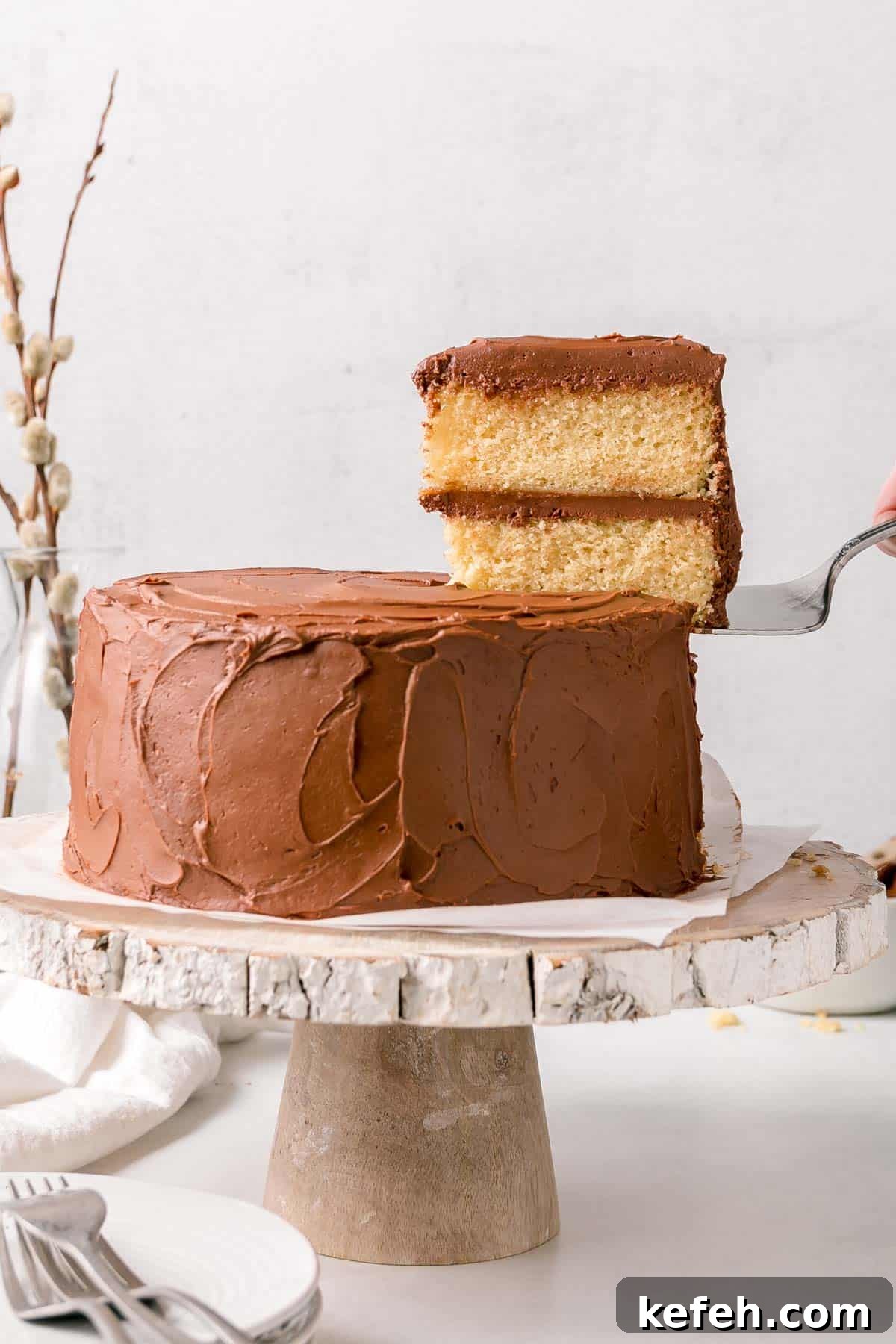 Two layer gluten free vanilla cake with chocolate frosting slice on serving spatula next to whole cake.