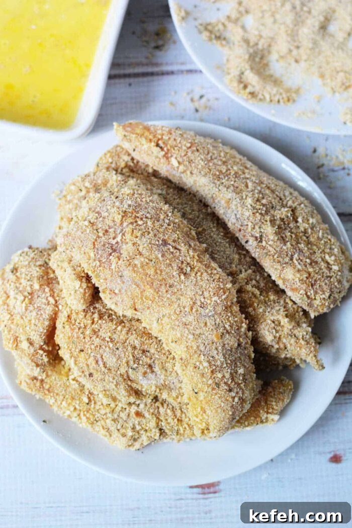 A white plate showcases crispy breaded chicken fillets, while in the backdrop, you can see all the essentials for creating your own masterpiece. This setup might just inspire your next cooking adventure after learning how to make chicken tenders at home.
