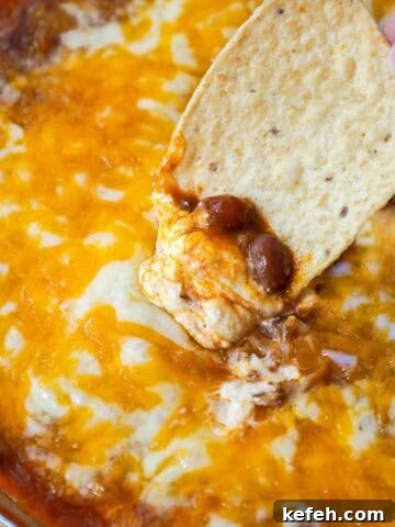 Tortilla chip scooping up hot chili cheese dip, revealing melted cheese, beans, and cream cheese.