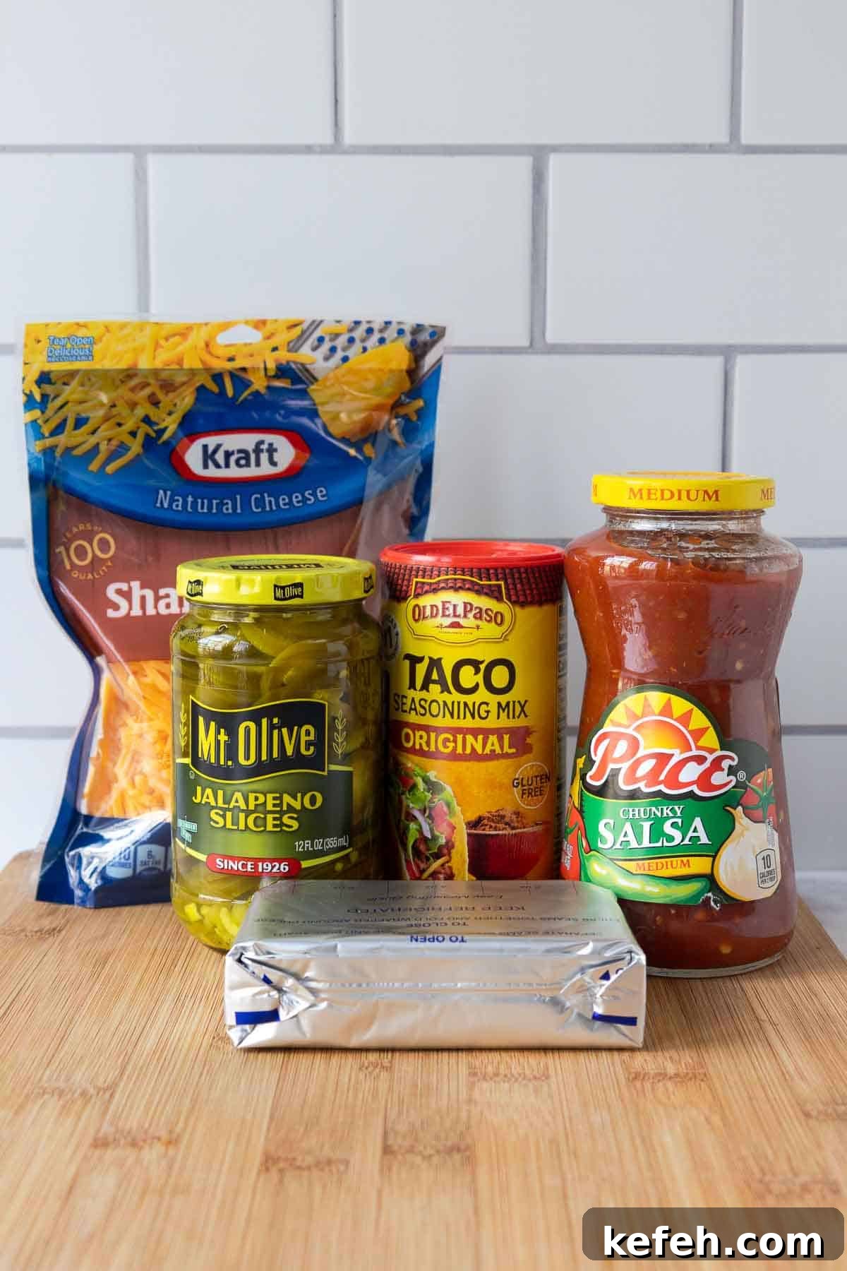Cream Cheese Salsa Dip Recipe for Parties and Game Day 3 Ingredients for cream cheese and salsa dip arranged on a wooden cutting board, including shredded cheddar cheese, jalapeño slices, taco seasoning, salsa, and a block of cream cheese.