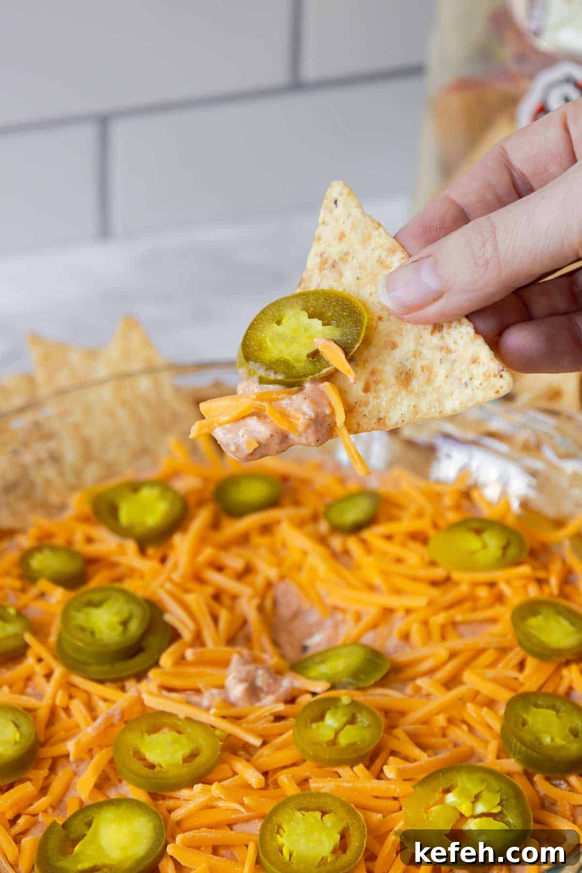 Cream Cheese Salsa Dip Recipe for Parties and Game Day 8 A hand holding a tortilla chip scooping up a bite of cream cheese and salsa dip with melted cheese and a jalapeño slice on top.