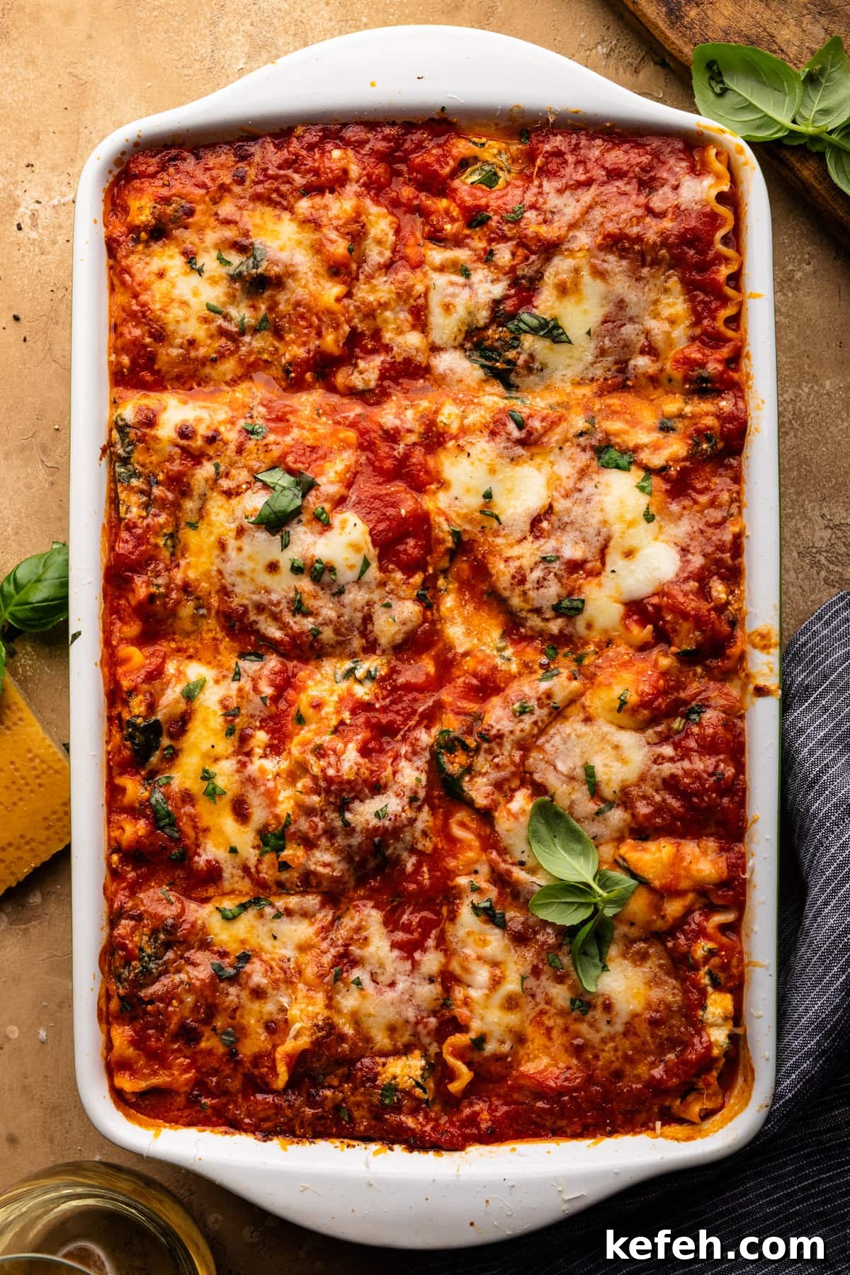 Creamy Classic Spinach Lasagna Recipe for Dinner 2 Lasagna in a white dish with green herbs sprinkled over it. The dish of lasagna is sitting on a tan colored background with a glass of white wine, a parmesan rind, and herbs arranged around it.
