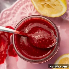 Closeup overhead of a small glass jar of homemade raspberry puree with a spoon in the top, on a pink decorative plate with lemon halves and raspberries.