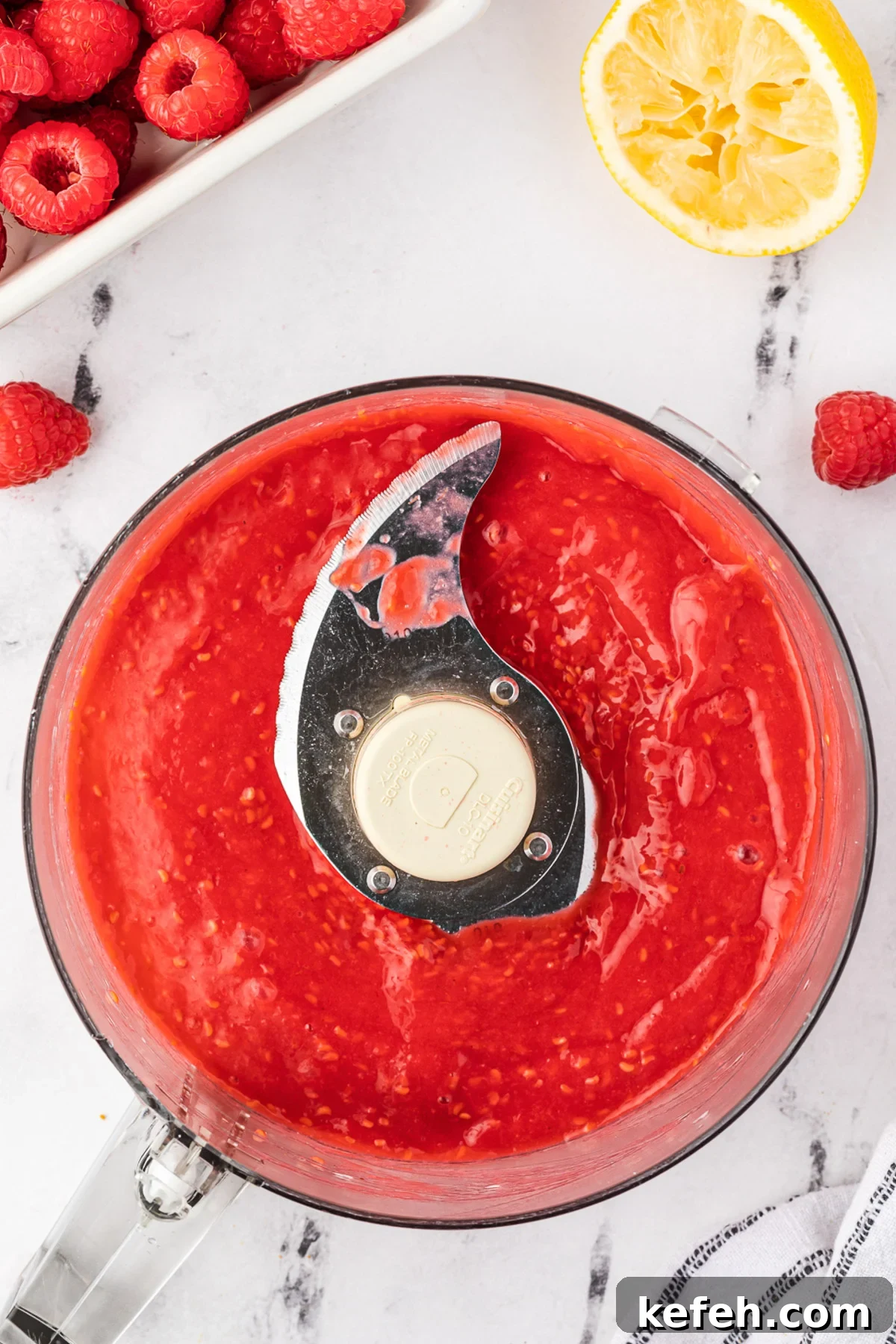 Overhead view of raspberry puree in a food processor bowl on a marble background with more raspberries and a tea towel next to it.