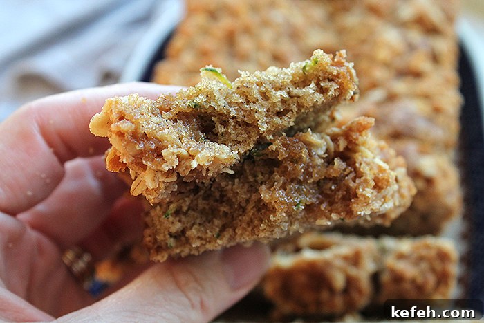 A slice of zucchini bread with streusel broken in half held above a sliced loaf of zucchini bread on an oval platter.