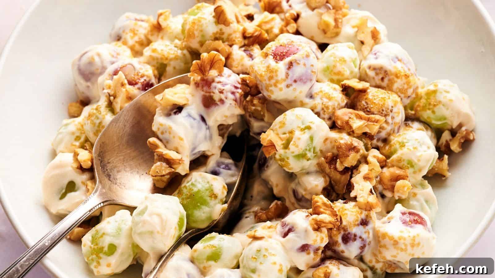 A bowl of grape salad with green and red grapes coated in creamy dressing, topped with chopped walnuts and brown sugar, with a spoon for serving.