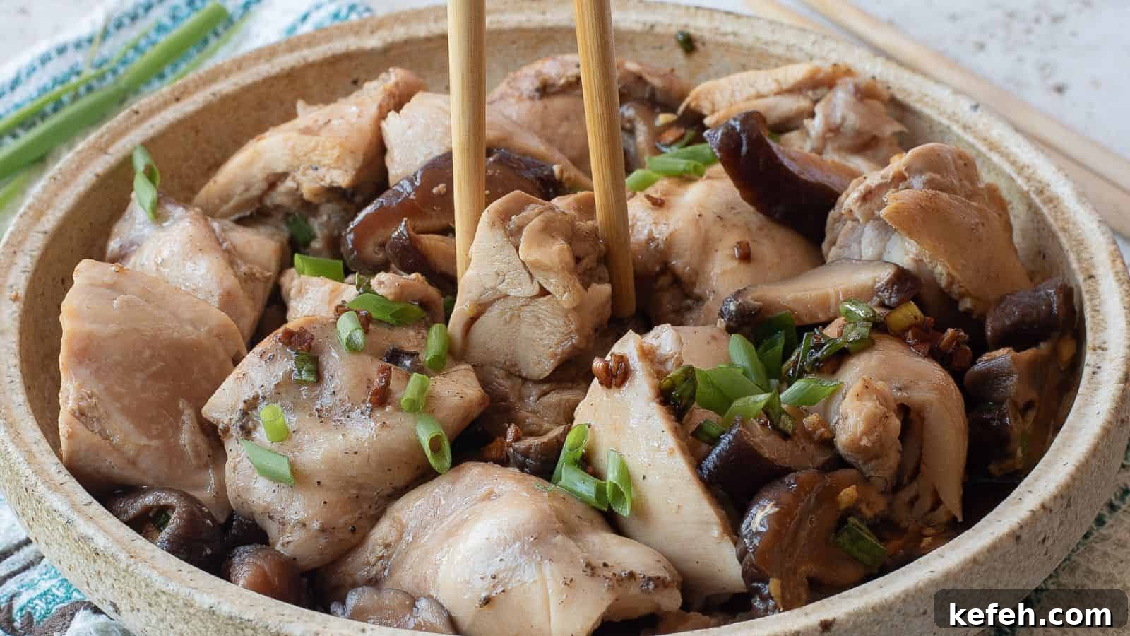 A bowl of steamed chicken pieces with mushrooms, garnished with chopped green onions, with chopsticks picking up a piece.