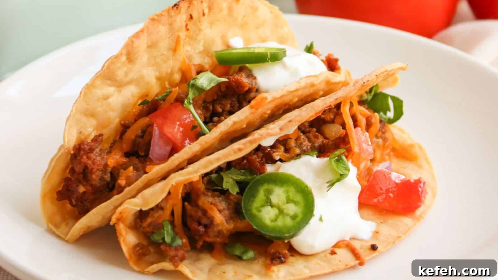 Two tacos with beef, diced tomatoes, jalapeño slices, shredded cheese, sour cream, and cilantro on a white plate.