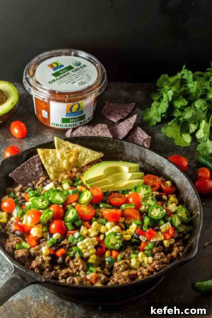 Crave-Worthy Quick Nachos: Customizable Recipes for Any Meal 6 Overhead shot of a cast iron skillet filled with healthy nachos made with seasoned ground beef, fresh vegetables, and avocado, with blue corn and yellow tortilla chips on the side.