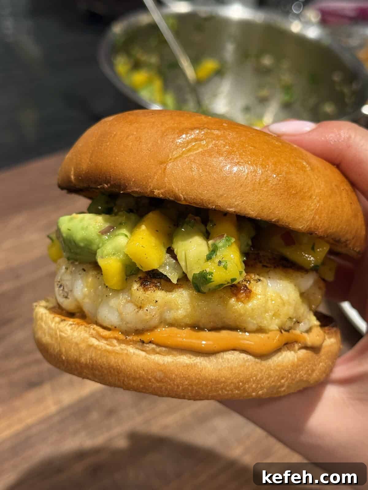 Shrimp Burgers with Mango Avocado Salad and Spicy Mayo 2 A hand holds a sandwich with a grilled patty, topped with a colorful mango and avocado salsa, and orange sauce on a soft, shiny bun. A bowl with more salsa is blurred in the background.