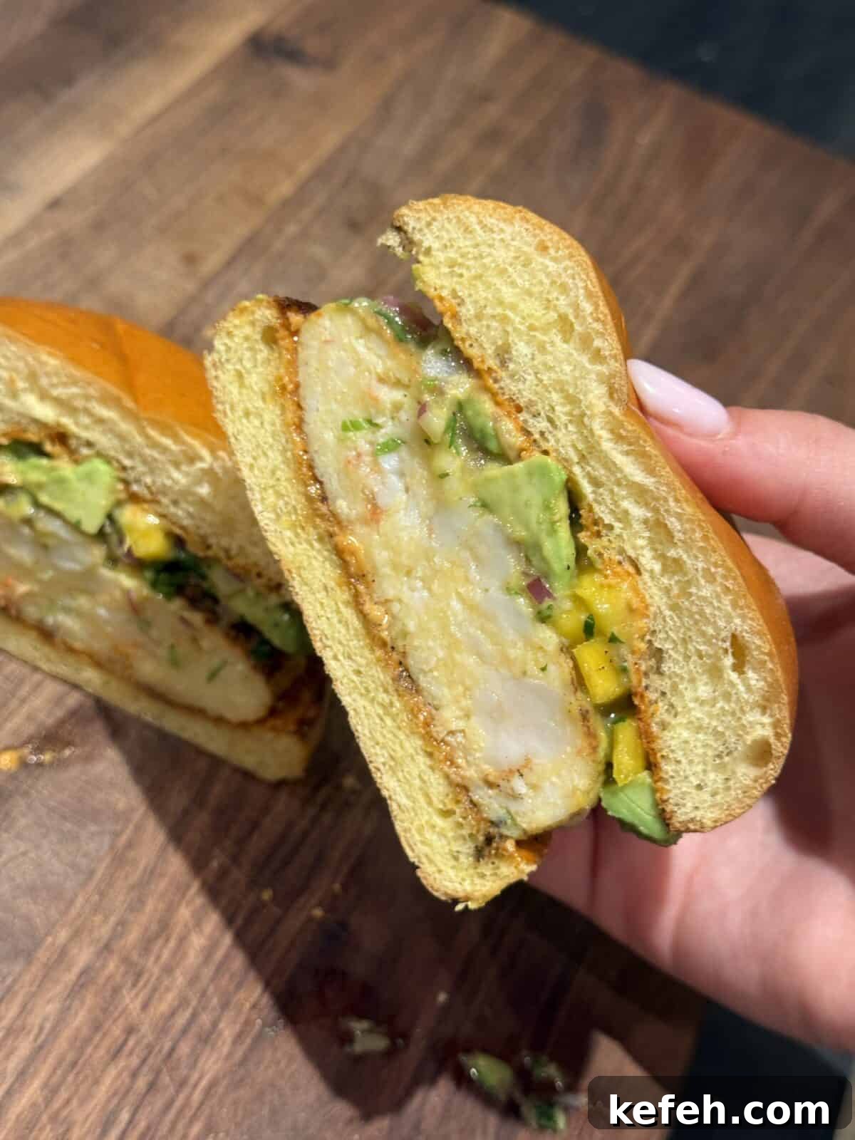 Shrimp Burgers with Mango Avocado Salad and Spicy Mayo 3 A hand holds half of a sandwich with a thick piece of breaded chicken, sliced avocado, chopped herbs, and a soft bun, shown on a wooden surface.