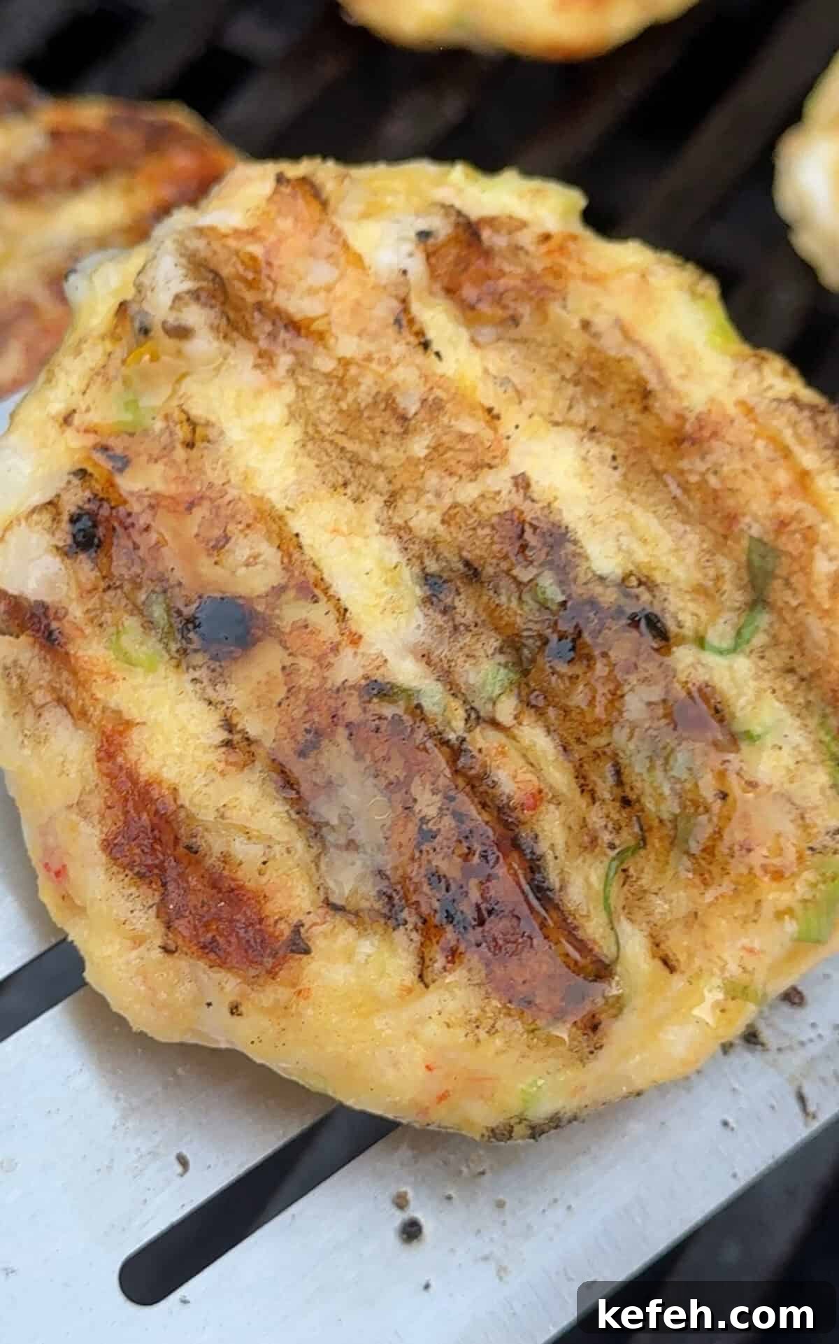 Shrimp Burgers with Mango Avocado Salad and Spicy Mayo 4 A close-up of a golden-brown grilled patty with visible grill marks, sitting on a metal spatula. The patty appears to contain small pieces of vegetables and herbs.