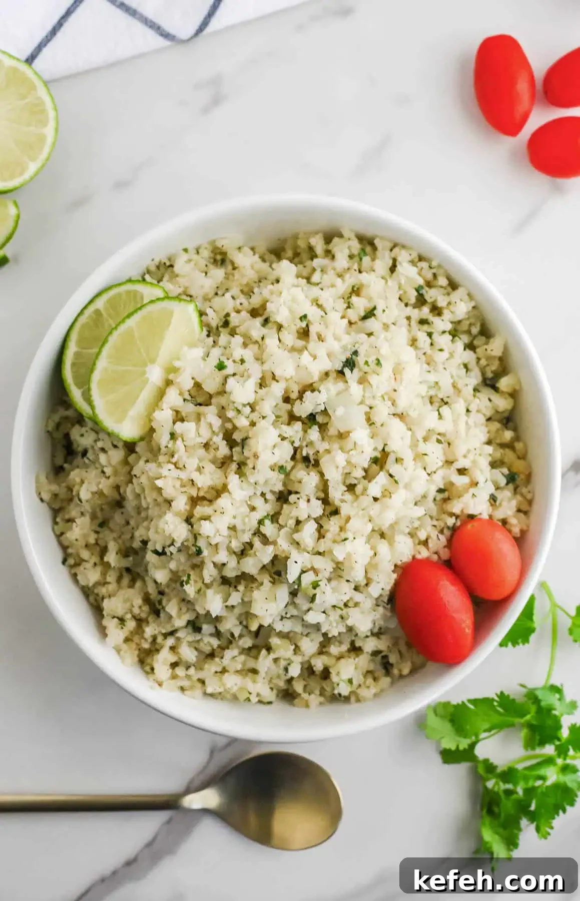 Cilantro Lime Cauliflower Rice Recipe for Flavorful Low-Carb Sides 11 A bowl of cilantro cauliflower lime rice with lime wedges and cherry tomatoes on a marble countertop.