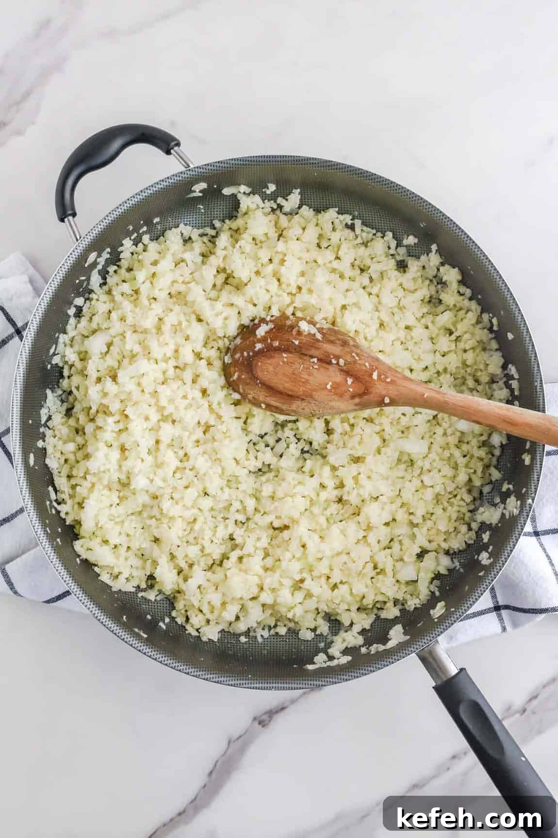 Cilantro Lime Cauliflower Rice Recipe for Flavorful Low-Carb Sides 6 A frying pan filled with cilantro lime cauliflower rice and a wooden spoon on a marble countertop.