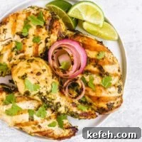 Four grilled cilantro lime chicken breasts with grilled onions and lime slices on a round plate.