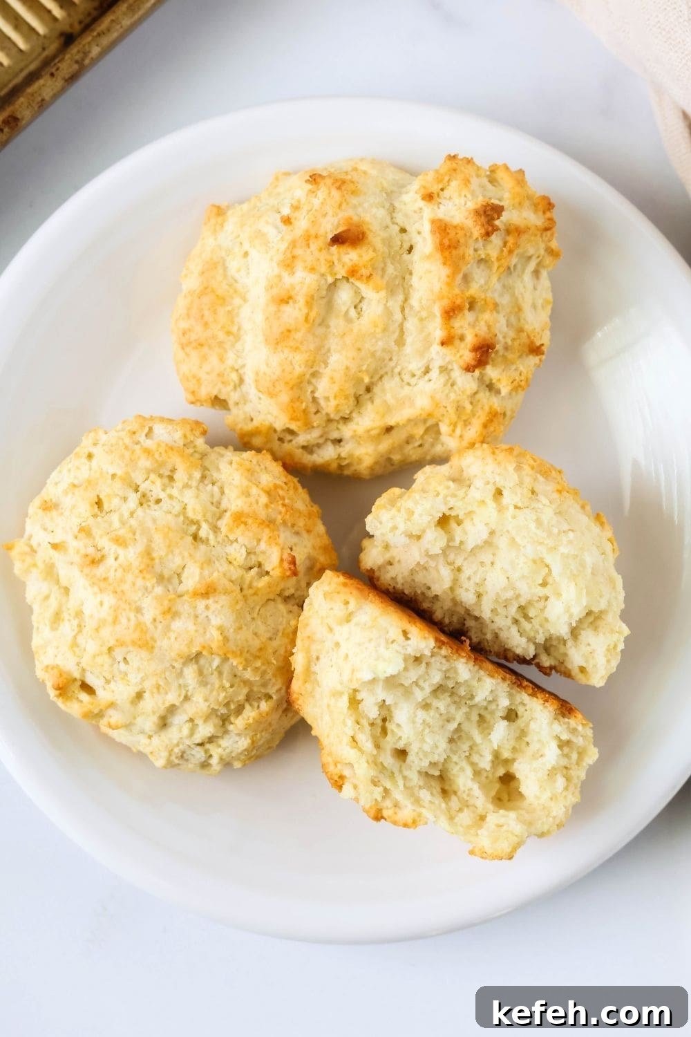 overhead view of three drop biscuits on a white plate. One biscuit is torn in half, showcasing the fluffy interior texture.