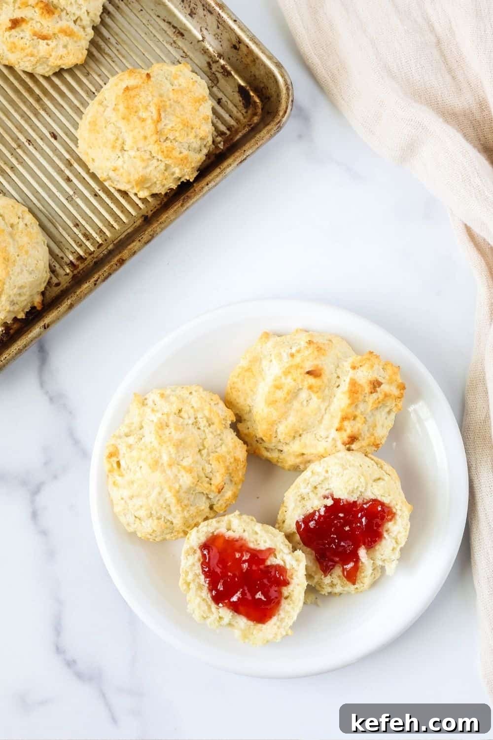 overhead view of a white plate with easy drop biscuits made with Bisquick on the plate. One biscuit is split in half and spread with jam. The pan of remaining biscuits is in the background.