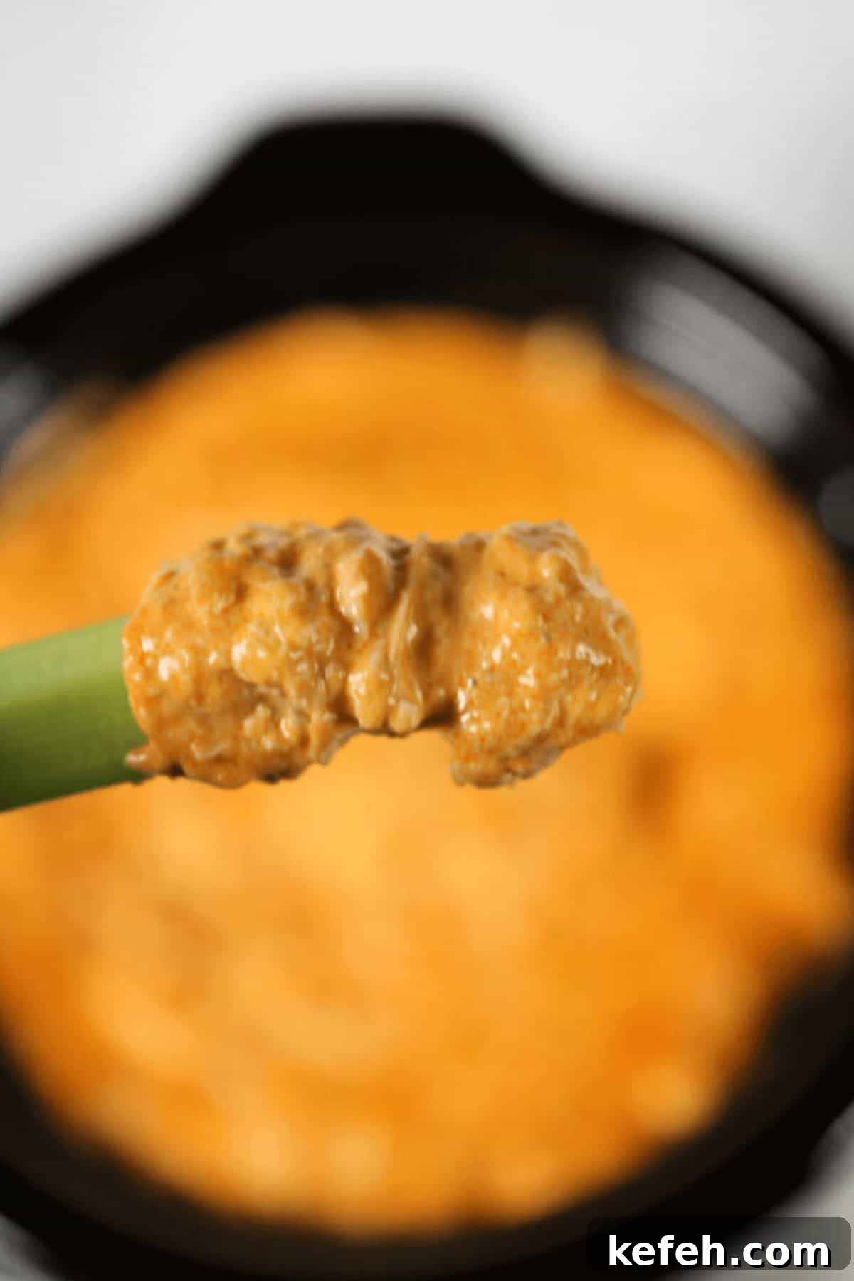 A close-up of a celery stick dipped, held above a dish brimming with more of the same tantalizing dip.