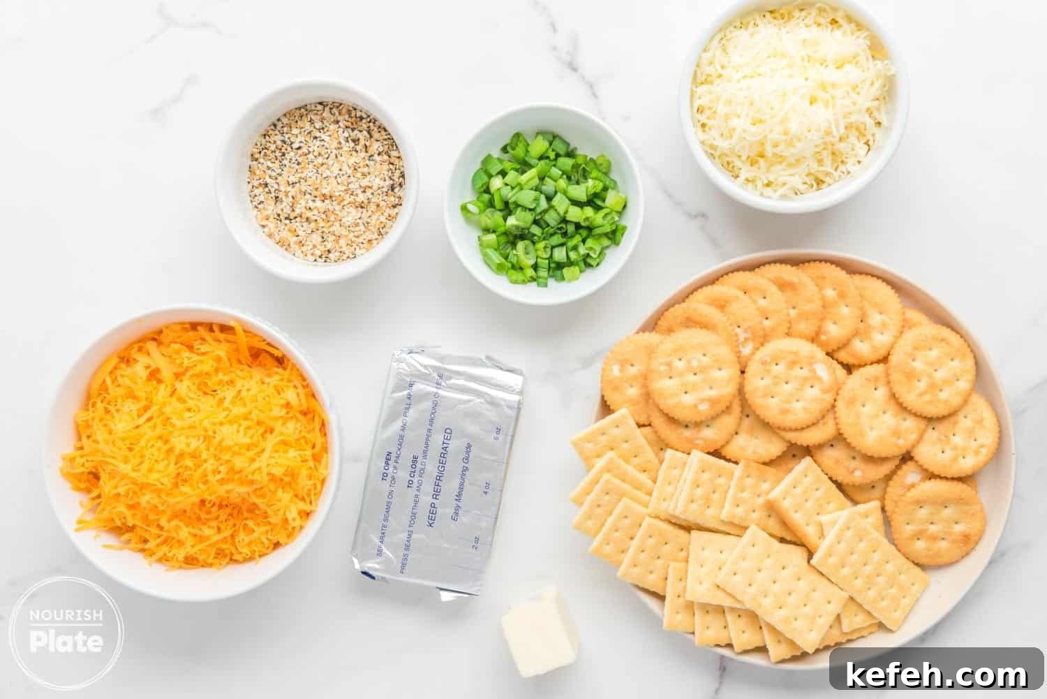 Everything Bagel Cheese Ball Recipe: Creamy Savory Appetizer 3 Overhead view of ingredients for Everything Bagel Cheese Ball, including shredded cheddar and colby jack cheeses, chopped scallions, Everything Bagel seasoning, a block of cream cheese, a small piece of butter, and a plate of Ritz and Club crackers on a white marble surface.