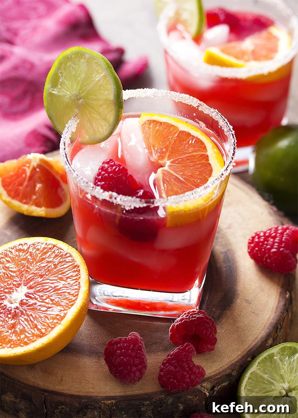 Two raspberry orange margaritas each garnished with fruit and in sugar rimmed glasses.