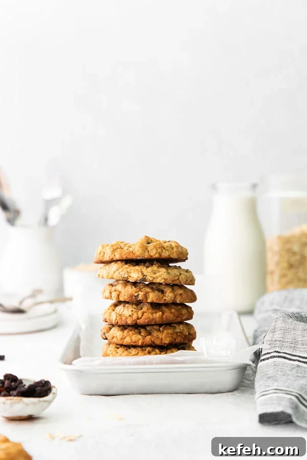 a stack of gluten free oatmeal raisin cookies piled high on a small silver metal sheet pan lined with white parchment paper with a glass of milk in the background