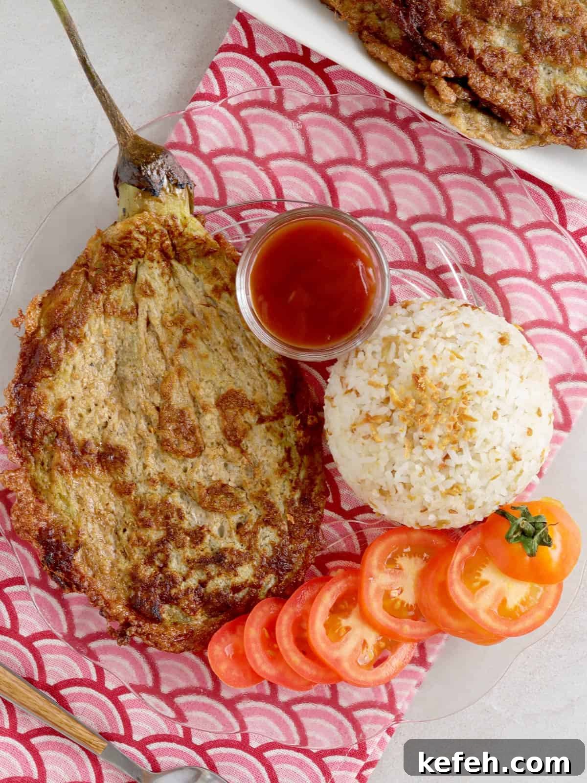 Tortang Talong on a clear glass plate with steamed rice and sliced tomatoes on the side.