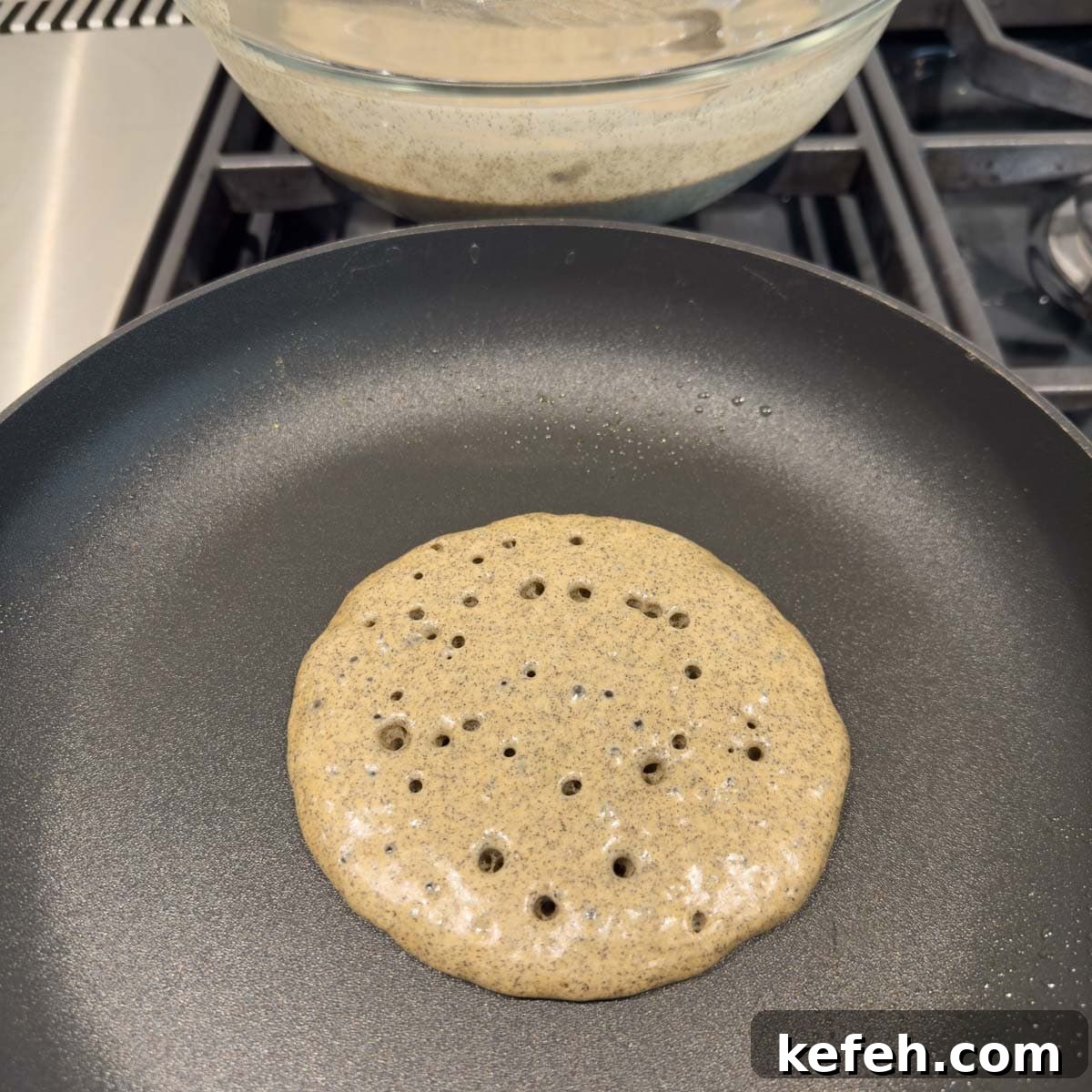 A pancake cooking in a non-stick pan, ready to flip with bubbles.