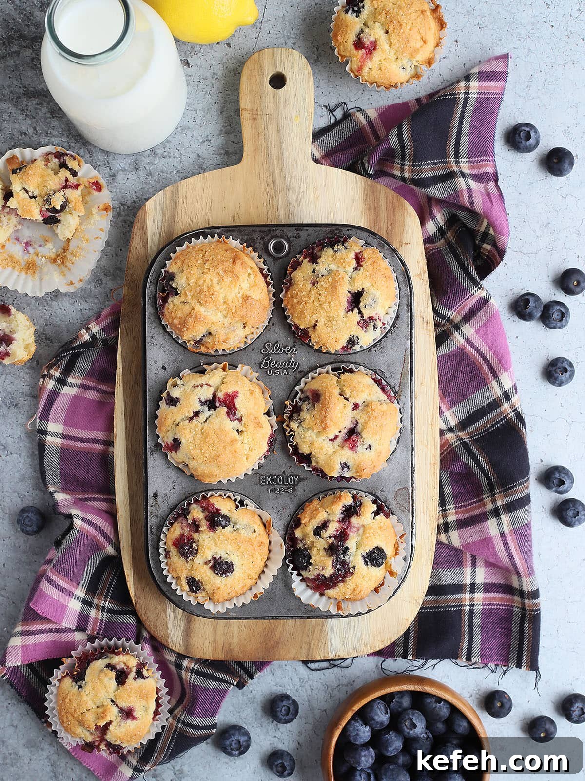 Buttermilk blueberry muffins in a muffin tin on a wooden cutting board with fresh blueberries and additional muffins on the side.