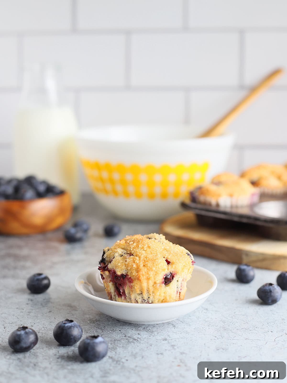 Single buttermilk blueberry muffin on a small dish with mixing bowls and baking ingredients in the background.