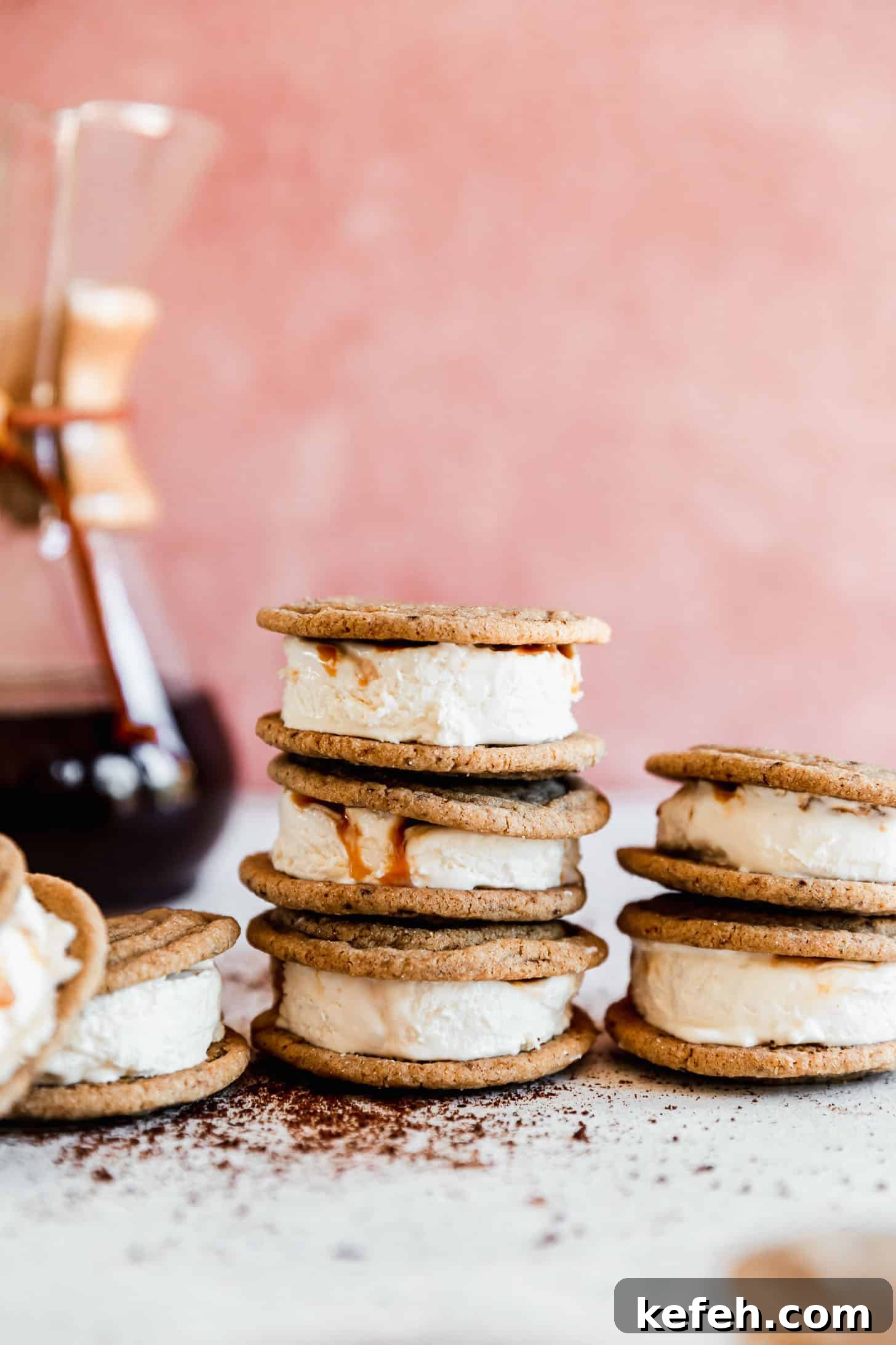 Salted Caramel Coffee Ice Cream Sandwiches Recipe 2 Coffee ice cream sandwiches with caramel swirl ice cream stacked on a white counter next to a Chemex in front of a pink background.