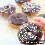Two fingers holding up a chocolate glazed protein donut with sprinkles with more donuts behind it.