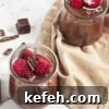 Two mason jars filled with chocolate pudding on a tan linen and topped with rapsberries and berries around them.