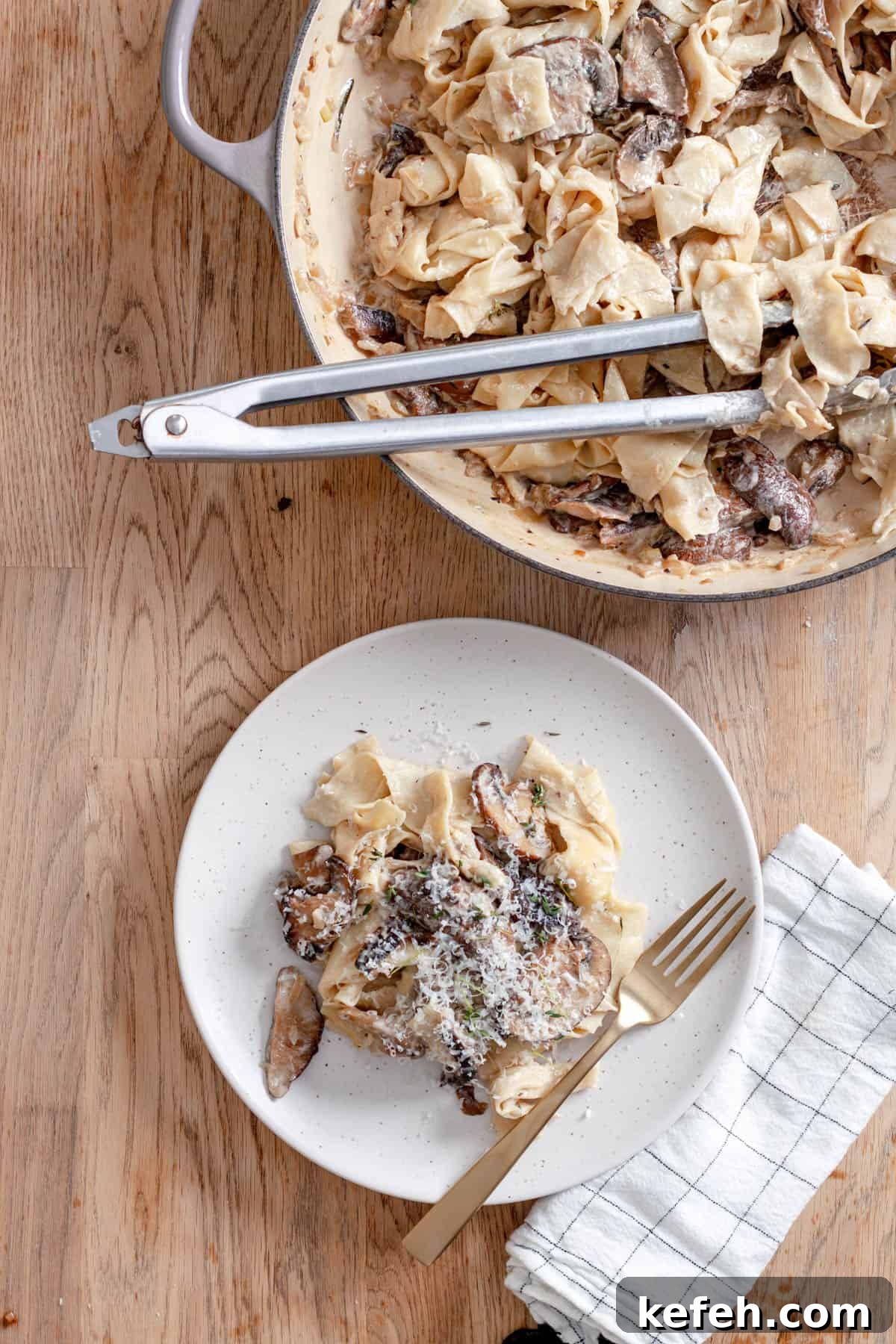 A serving of mushroom pappardelle on a white plate with a large skillet of more noodles to the side.