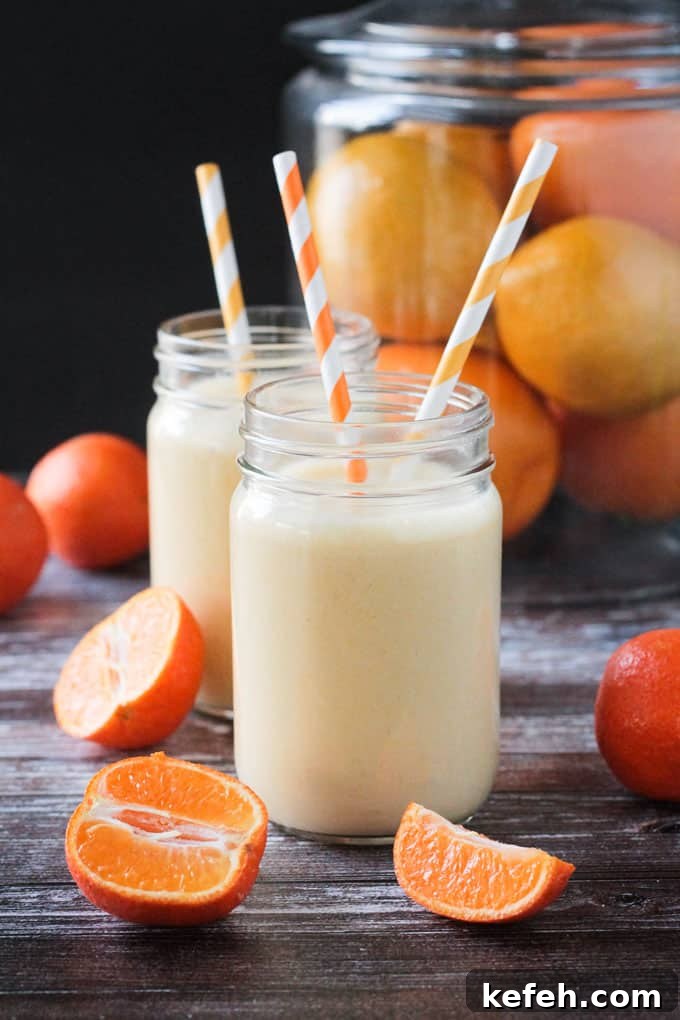 Two glass of clementine smoothie wit orange/white straws. Halved clementines rest nearby.