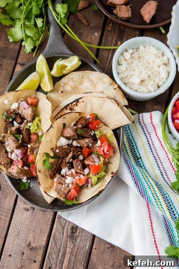 A plate of two delicious carne asada tacos with two tortillas and three slices of lime with a small white bowl of cheese on the side.