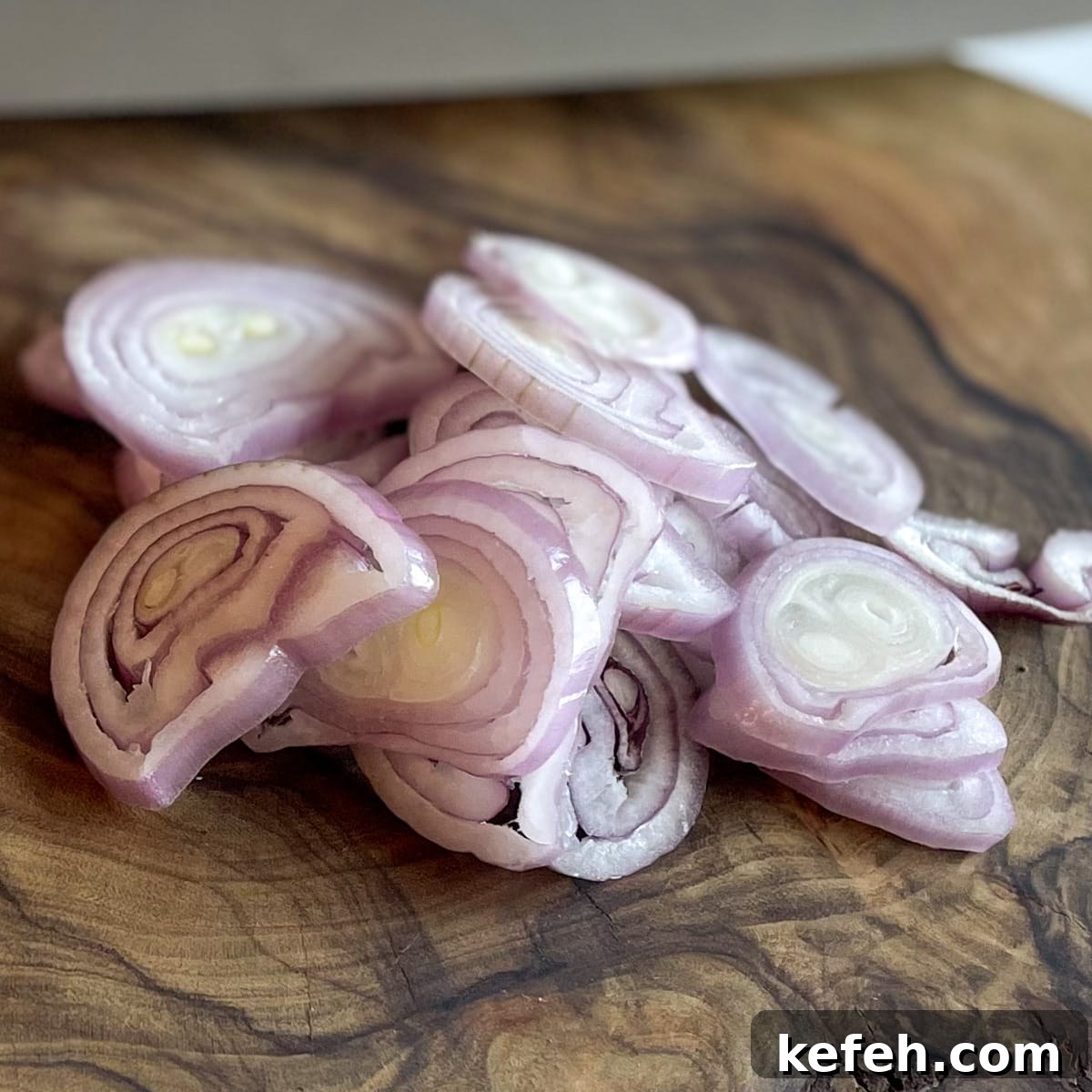 Rings of sliced shallot sit on a wooden cutting board with a knife blade in the background.