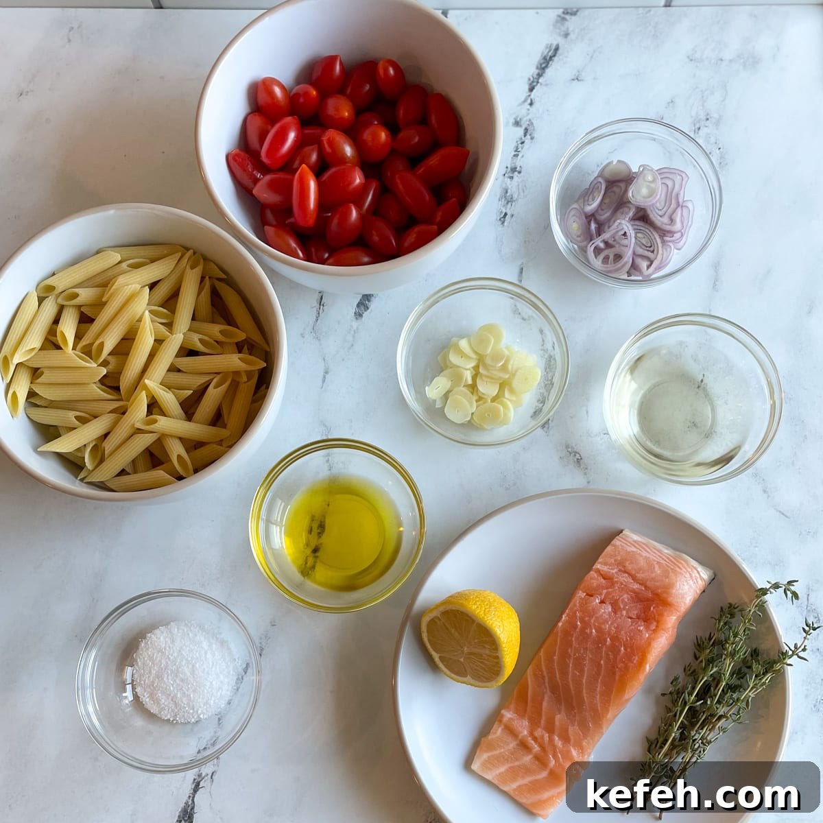 White bowls with penne pasta and cherry tomatoes, a white plate with a fillet of salmon, half of a lemon, and a bunch of thyme, and clear pinch bowls with salt, olive oil, sliced garlic, sliced shallots, and white wine sit on a white marble countertop.