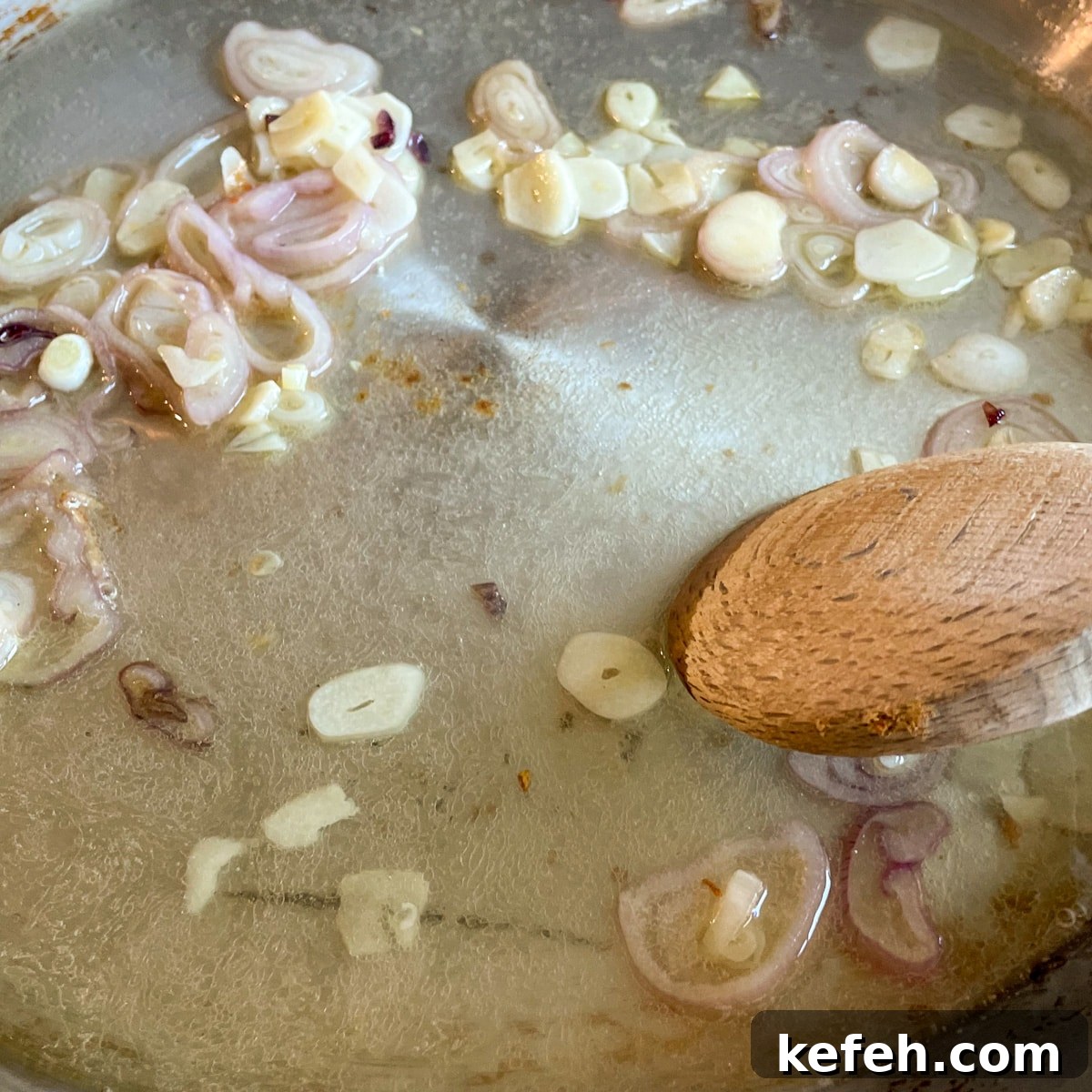 Sautéed shallots and garlic are shown in a stainless steel pan with white wine and lemon juice with a wooden spoon mixing the ingredients together.