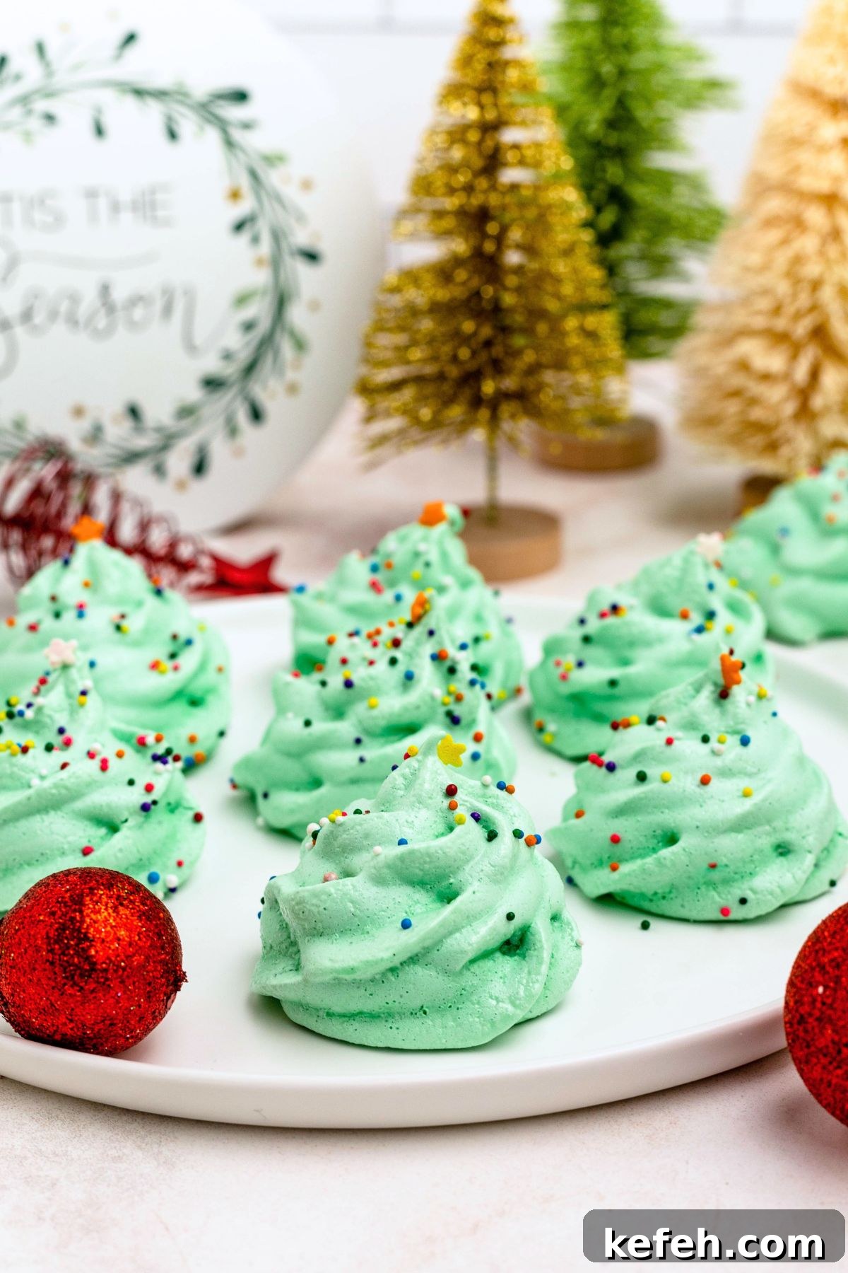 Green meringue trees on a white plate, with Christmas ornaments and mini trees on the table. 