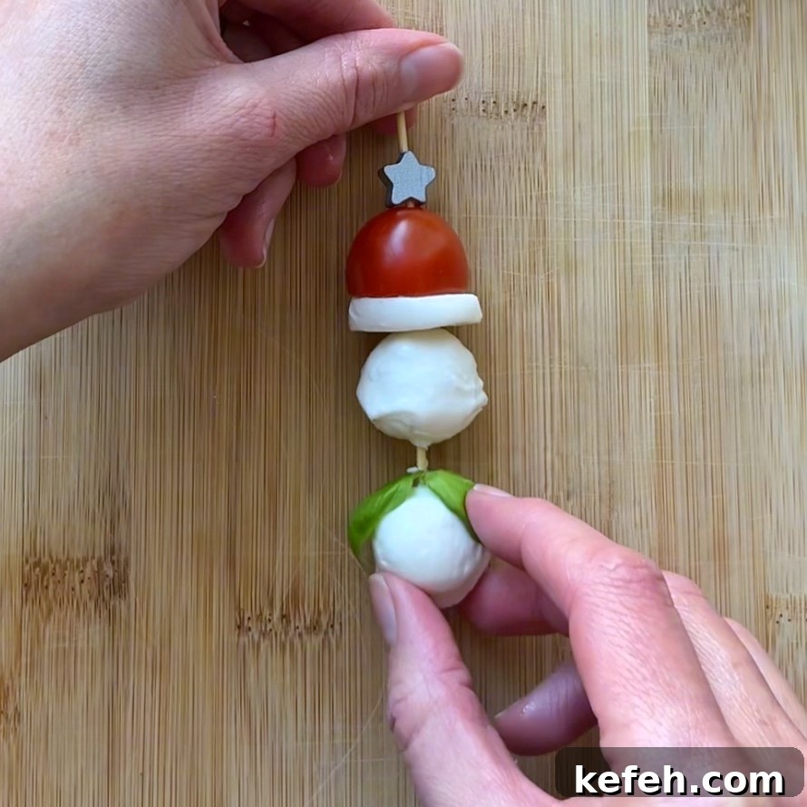 Christmas Party Appetizer Ideas to Impress Your Guests 5 Hands assembling a skewer with a cherry tomato, mozzarella balls, a basil leaf, and a slice of mozzarella cheese on a wooden cutting board.