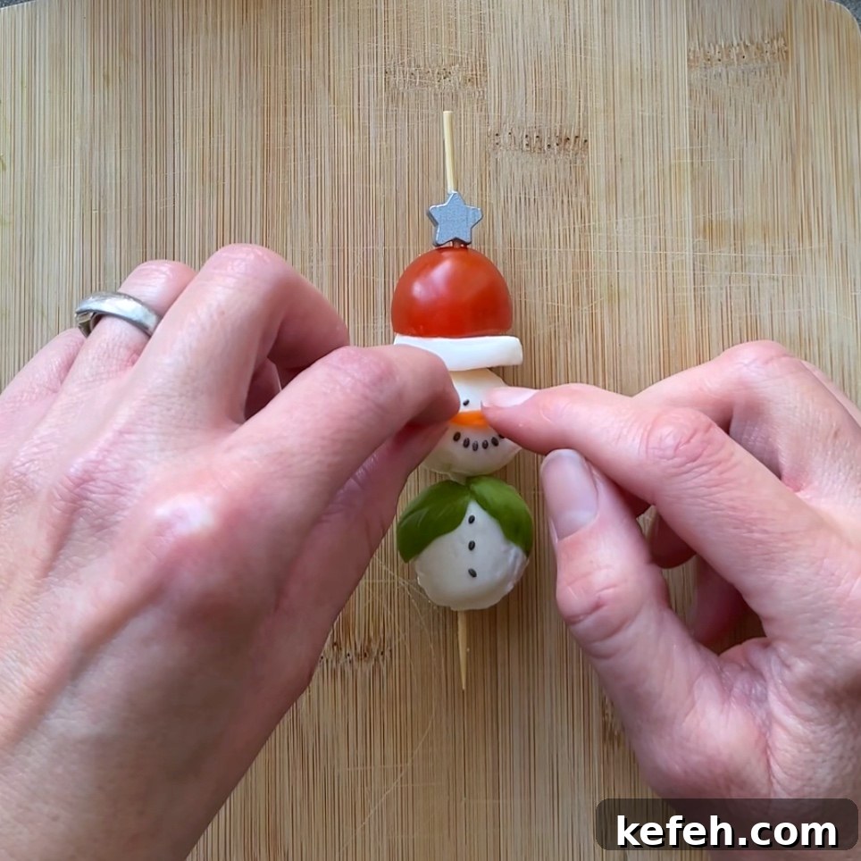 Christmas Party Appetizer Ideas to Impress Your Guests 6 Two hands assemble a snowman-shaped snack on a wooden board using a skewer with tomato, mozzarella, and basil.