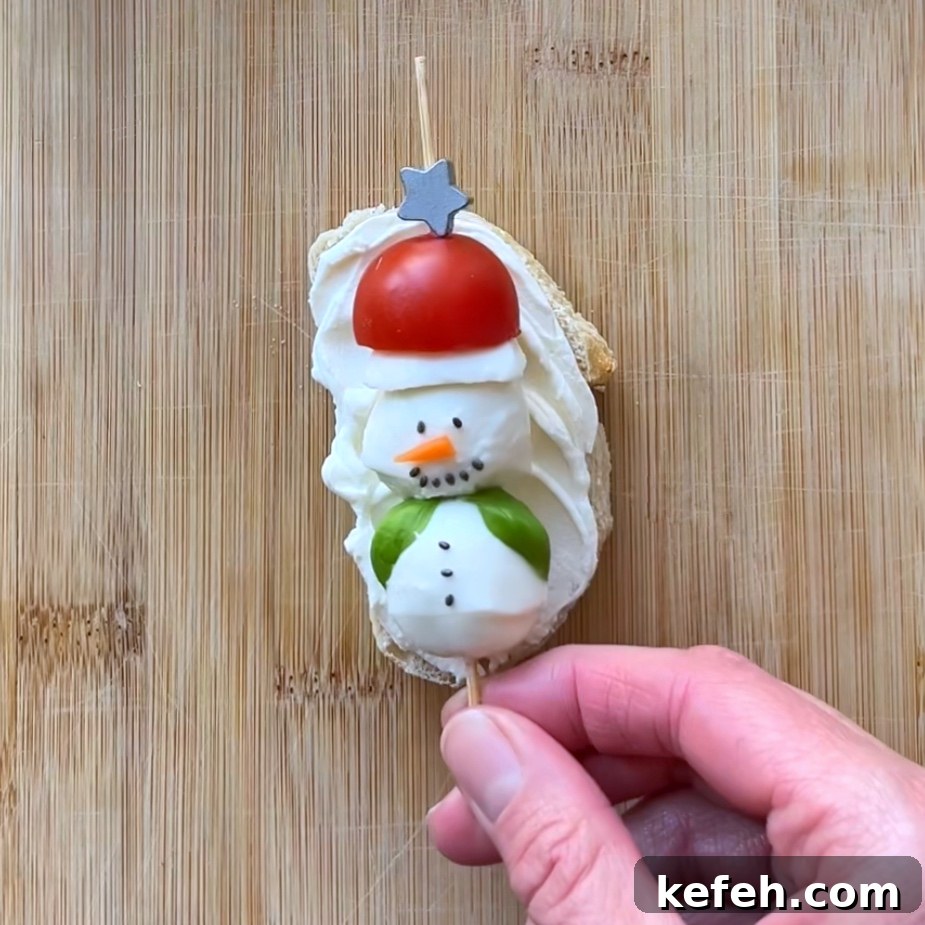 Christmas Party Appetizer Ideas to Impress Your Guests 7 A hand holds a skewer with a snowman made of mozzarella, tomato, and basil on cream cheese–spread bread.