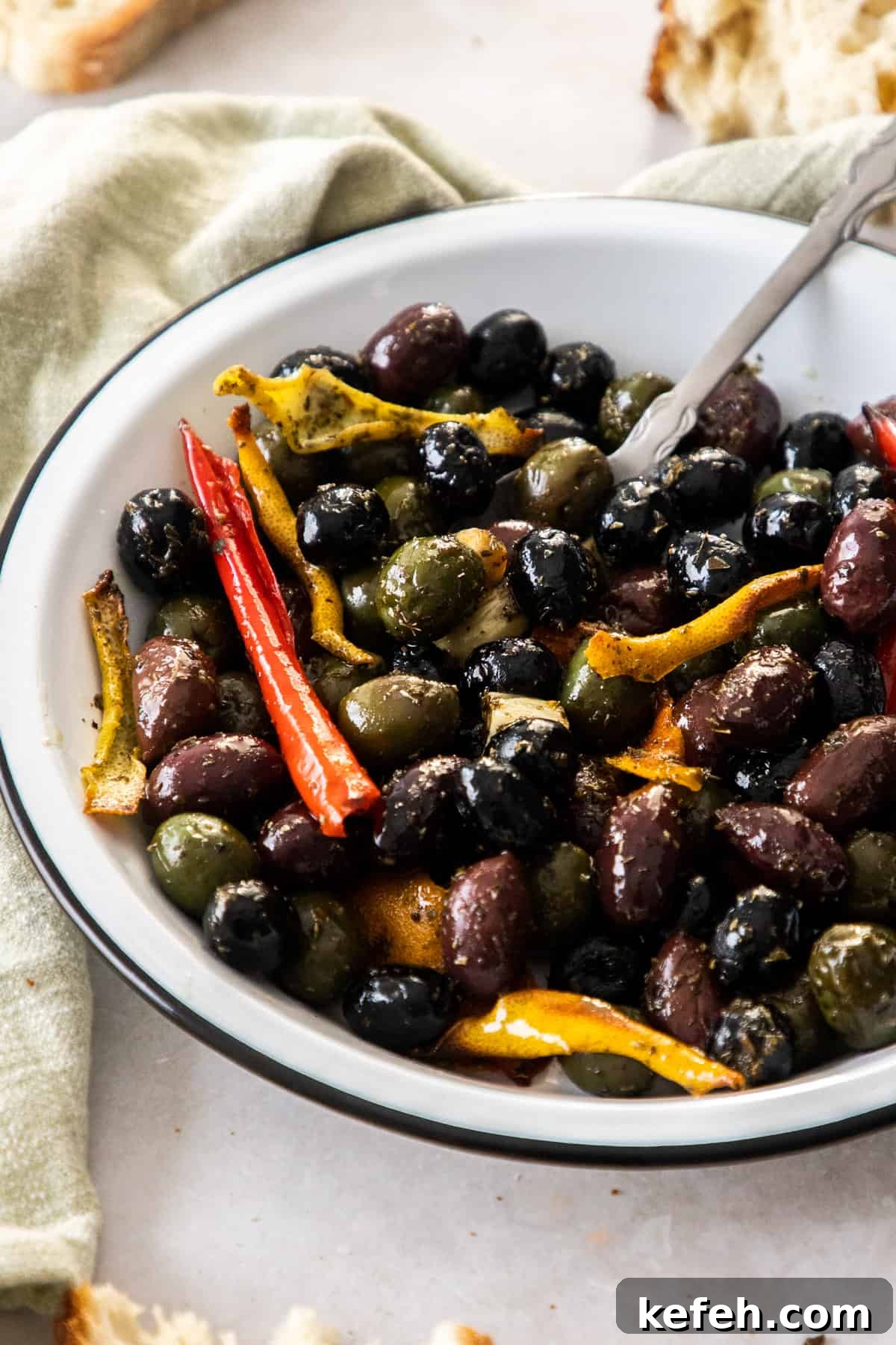 Round white dish with roasted olives, and a silver spoon sitting in it, and a green towel and crusts of bread around the edge.