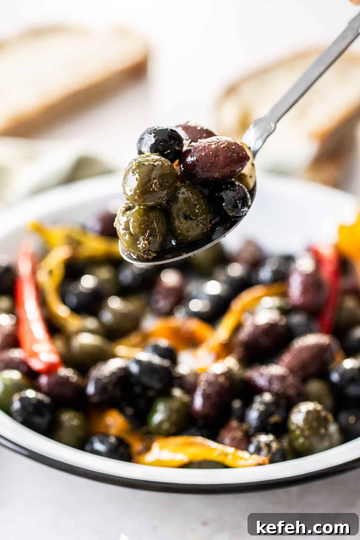 Spoon holding up a scoop of roasted olives, with the dish of olives in the background.
