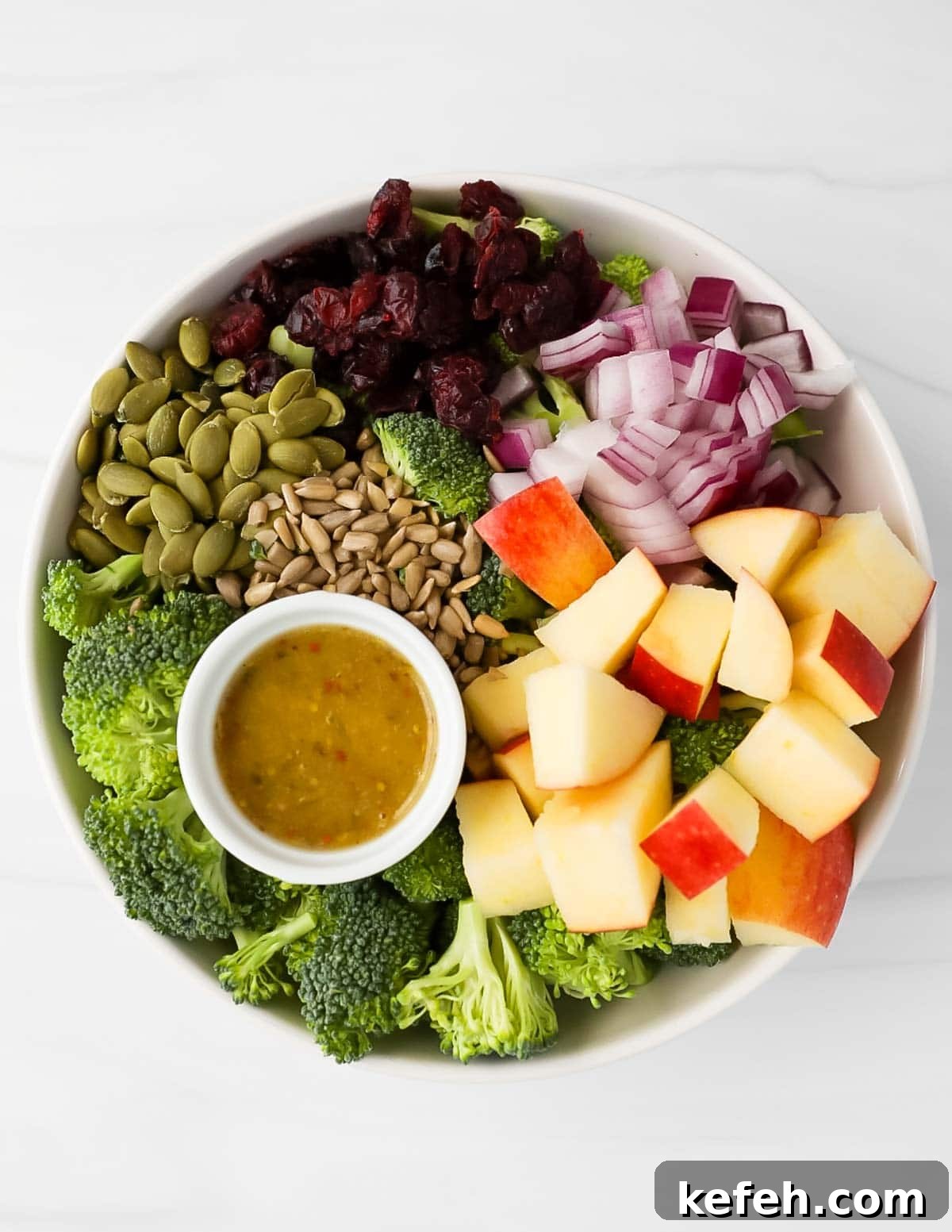 Crispy Broccoli Crunch Salad with Bacon and Sunflower Seeds 4 A large white bowl filled with broccoli, apples, red onion, dried cranberries, seeds, and a dish filled with dressing.